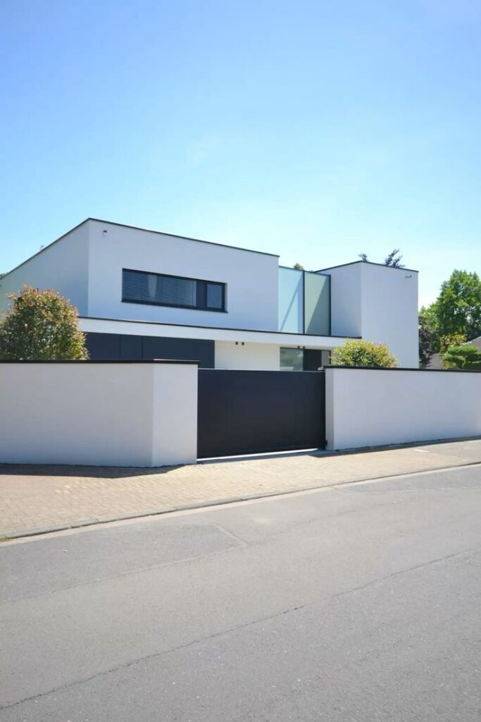 Modern white house with minimalist design, flat roof, and sleek windows, surrounded by a white wall on a sunny day.