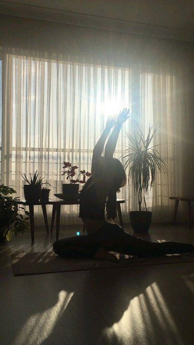 Person practicing yoga in a sunlit room with plants, performing a split and reaching upward.