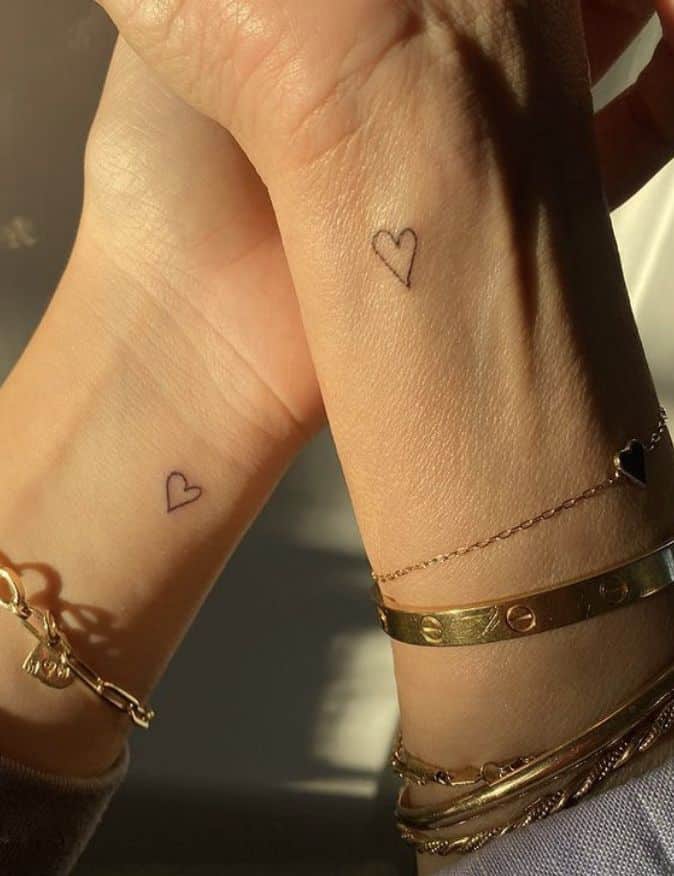Minimalist heart tattoos on wrists with gold bracelets in sunlight.