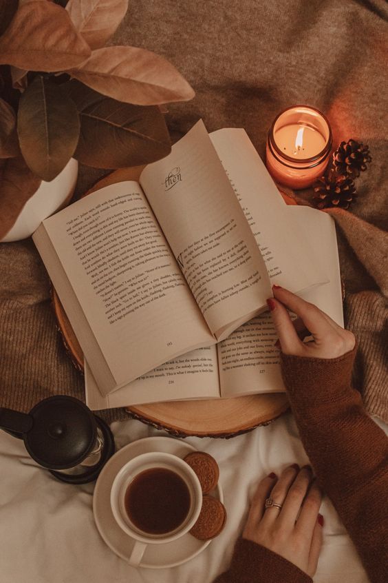 Cozy reading setup with open book, lit candle, coffee cup, and cookies on a warm blanket. Relaxing autumn atmosphere.