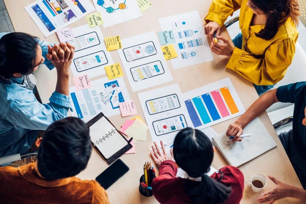 Team brainstorming on mobile UI/UX design with colorful charts and sketches spread on a table.