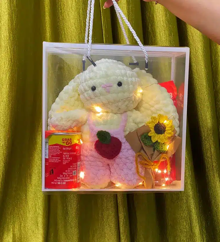 Hand holding gift box with plush bunny, snacks, and knitted sunflower against gold curtain.