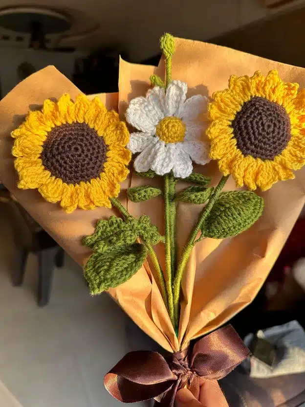 Crochet sunflowers and daisy bouquet wrapped in brown paper with a brown ribbon, captured in warm sunlight.