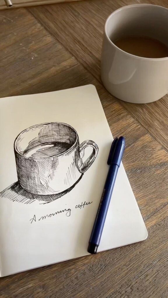Illustrated coffee cup in notebook with pen and real mug on wooden table, morning inspiration.