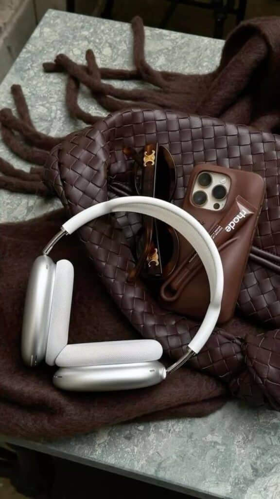 Silver headphones, sunglasses, phone in brown case on woven bag, stylish accessories on a textured surface.