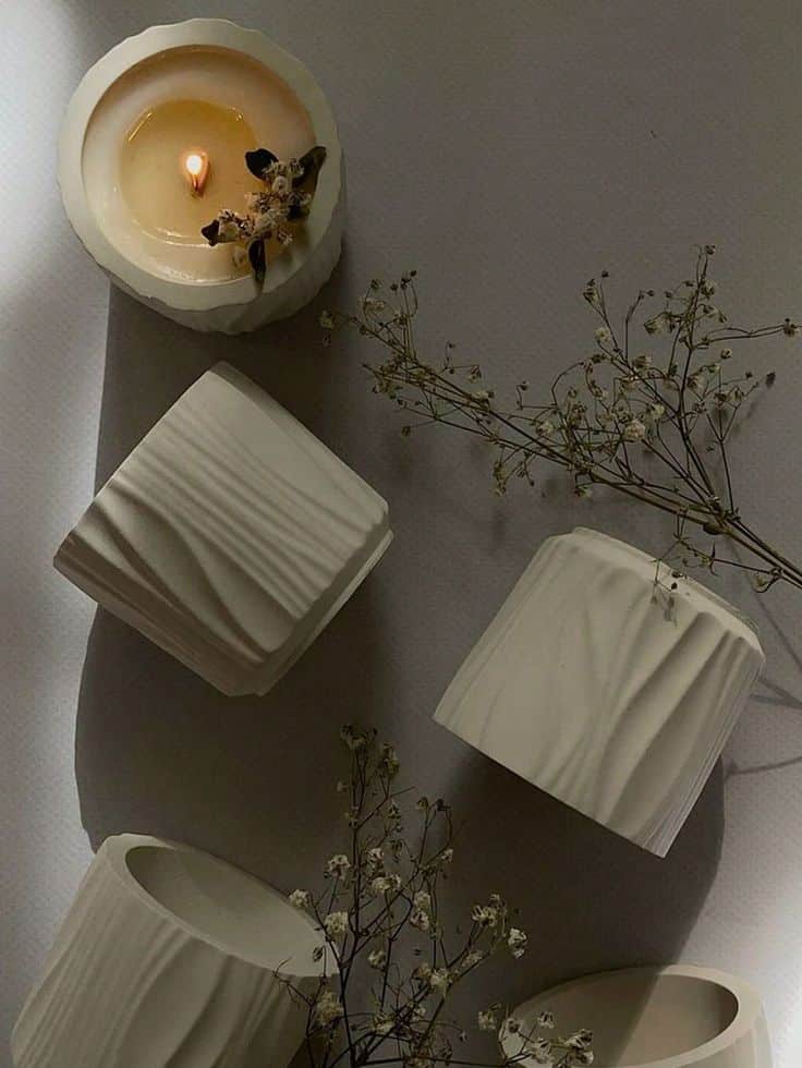Lit candle with floral decor, surrounded by white ceramic holders and delicate dried flowers on a textured surface.