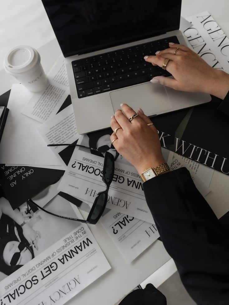 Person working on laptop amidst stylish magazines and coffee, emphasizing modern work environment and design.