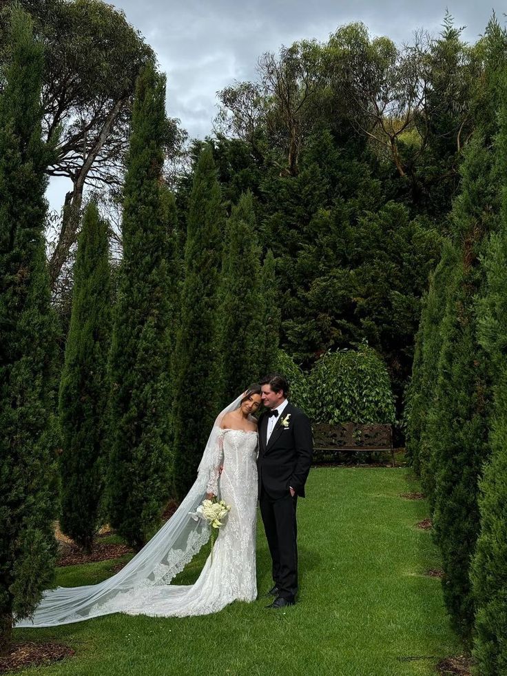 Bride and groom embrace in a lush garden, surrounded by tall green trees, under a cloudy sky.