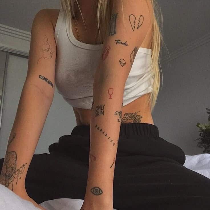 Woman in a white crop top with multiple minimalist tattoos on arms and torso, leaning forward with black pants.