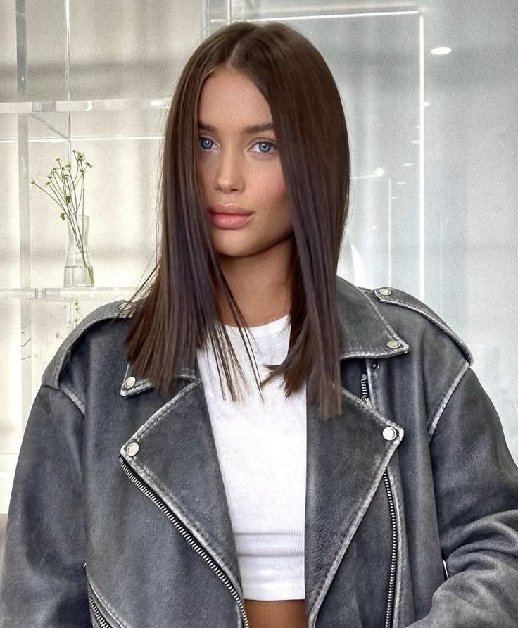 Fashionable woman in gray leather jacket and white top, with straight brown hair and blue eyes in a modern setting.
