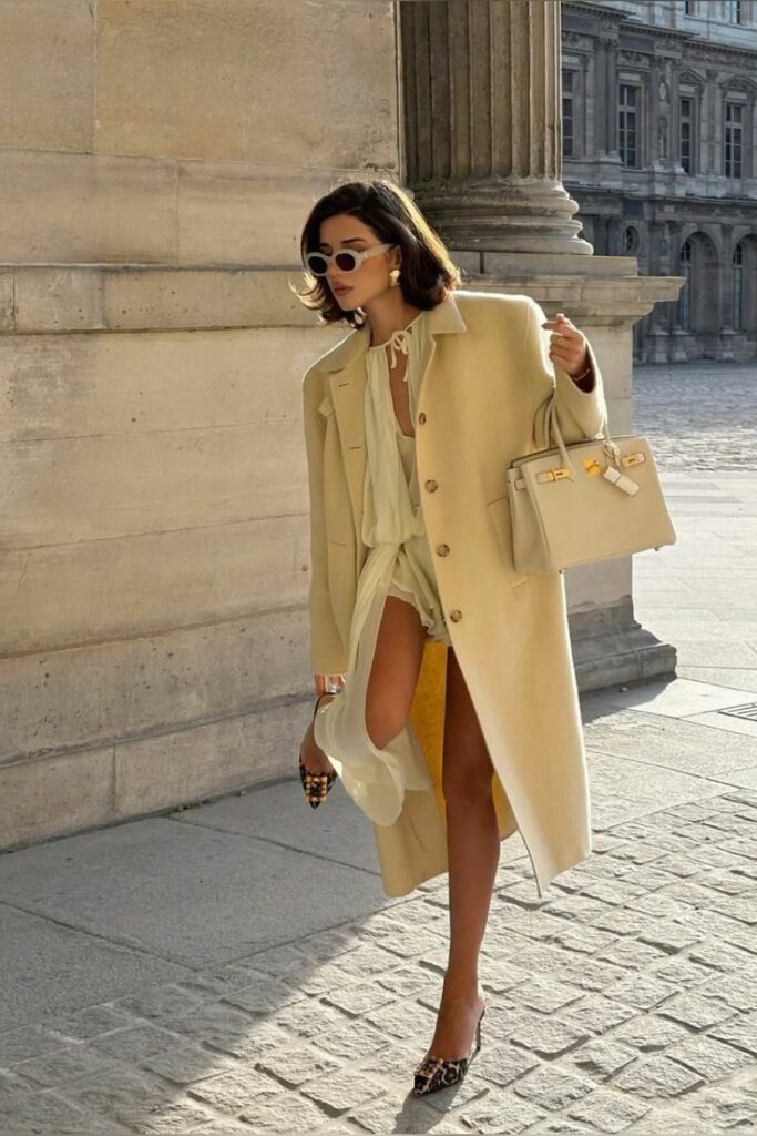 _ 2 Stylish woman in creamy coat and sunglasses, holding a designer bag, struts confidently by historic building. | Sky Rye Design Stylish woman in creamy coat and sunglasses, holding a designer bag, struts confidently by historic building.