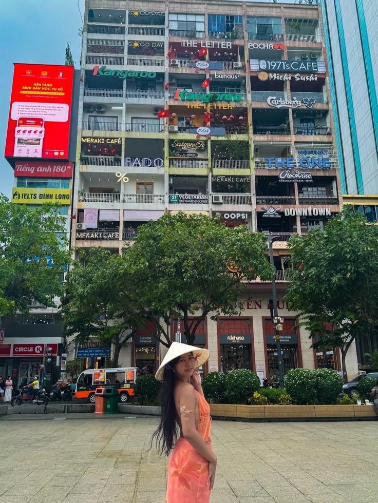 Woman in orange dress and conical hat in front of colorful building with café signs in a bustling city.
