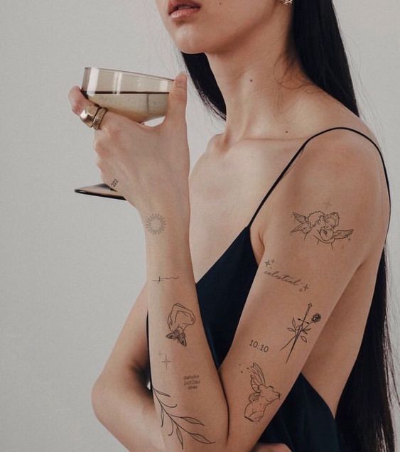 Woman with minimalist tattoos on arms holding a glass, wearing a black dress.