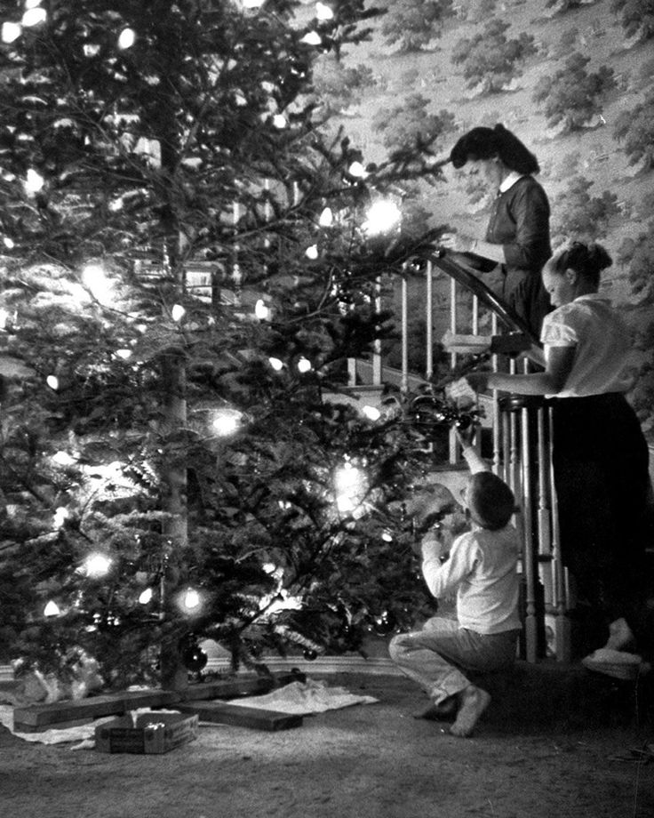 Vintage Christmas scene with three people decorating a large tree with lights and ornaments, creating a festive atmosphere.