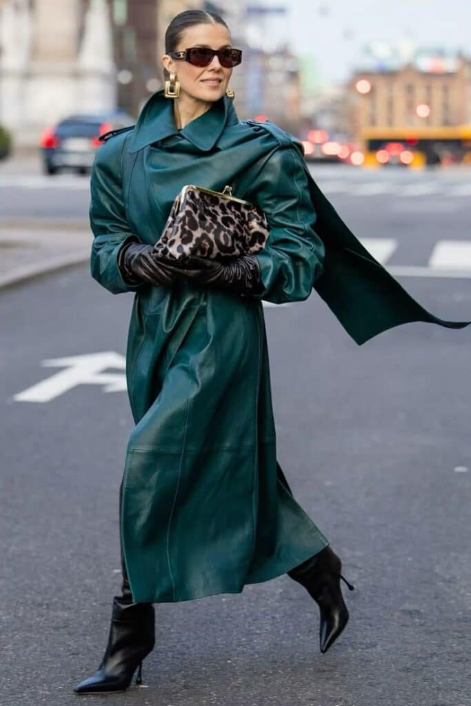 Fashionable woman in teal trench coat, sunglasses, stylish black boots, holding a leopard print clutch, city street.