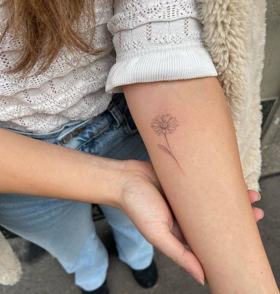 Delicate daisy tattoo on forearm, woman in white sweater and jeans, showcasing minimalist ink art.