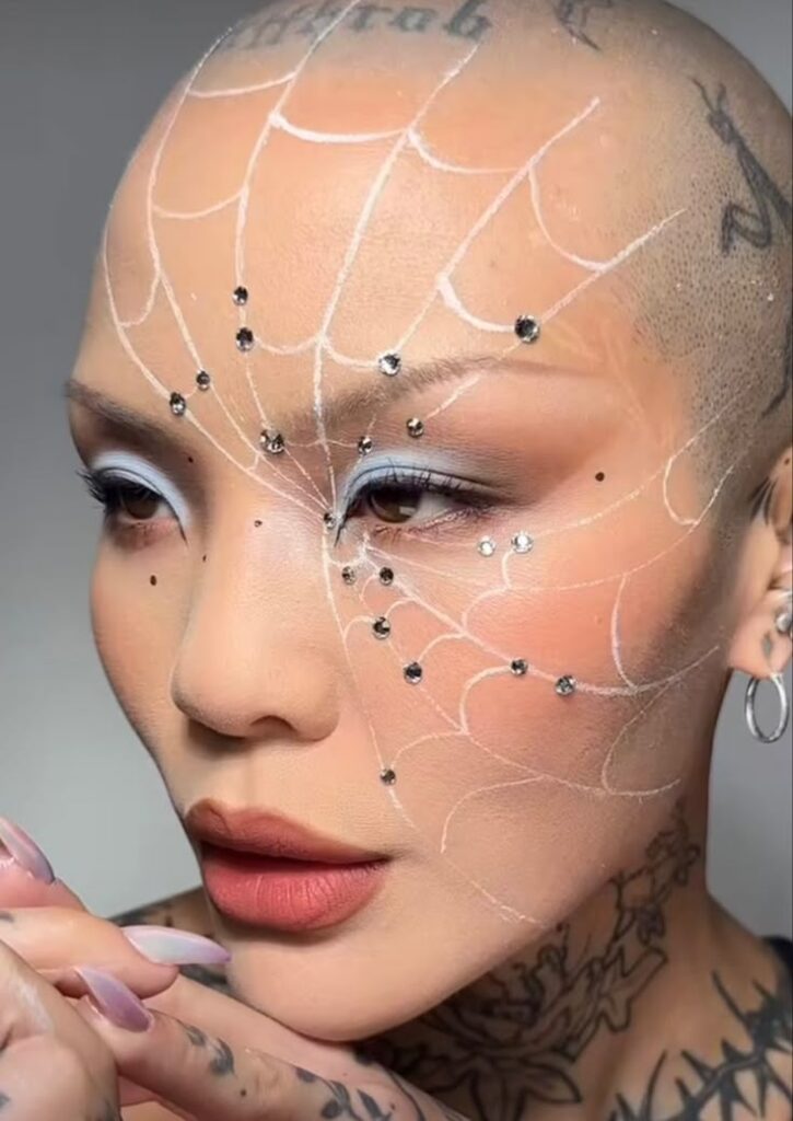 Bald woman with artistic web makeup and facial gems, focusing on elegant bold eye look and tattoos.
