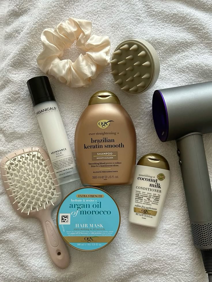 Hair care essentials featuring shampoos, conditioner, hair mask, brush, scrunchie, massager, and hairdryer on a towel.