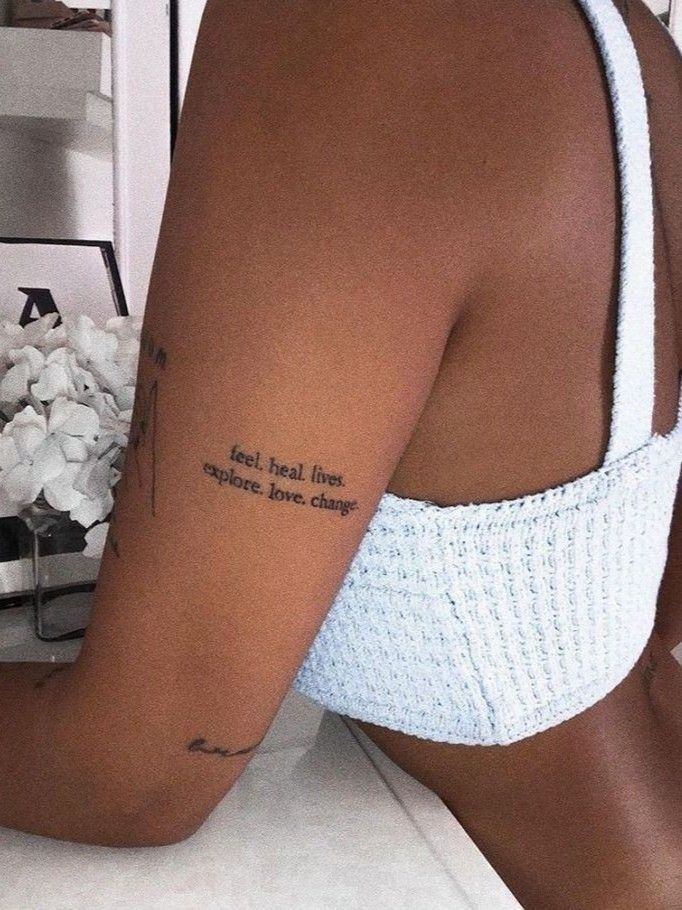Woman's arm tattoo with inspiring words, wearing a blue textured top, next to white flowers.