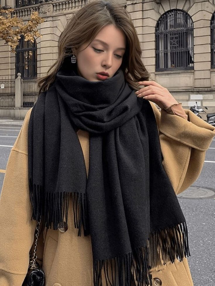 Woman in a tan coat with a black scarf, standing in a city street with a historic building in the background.