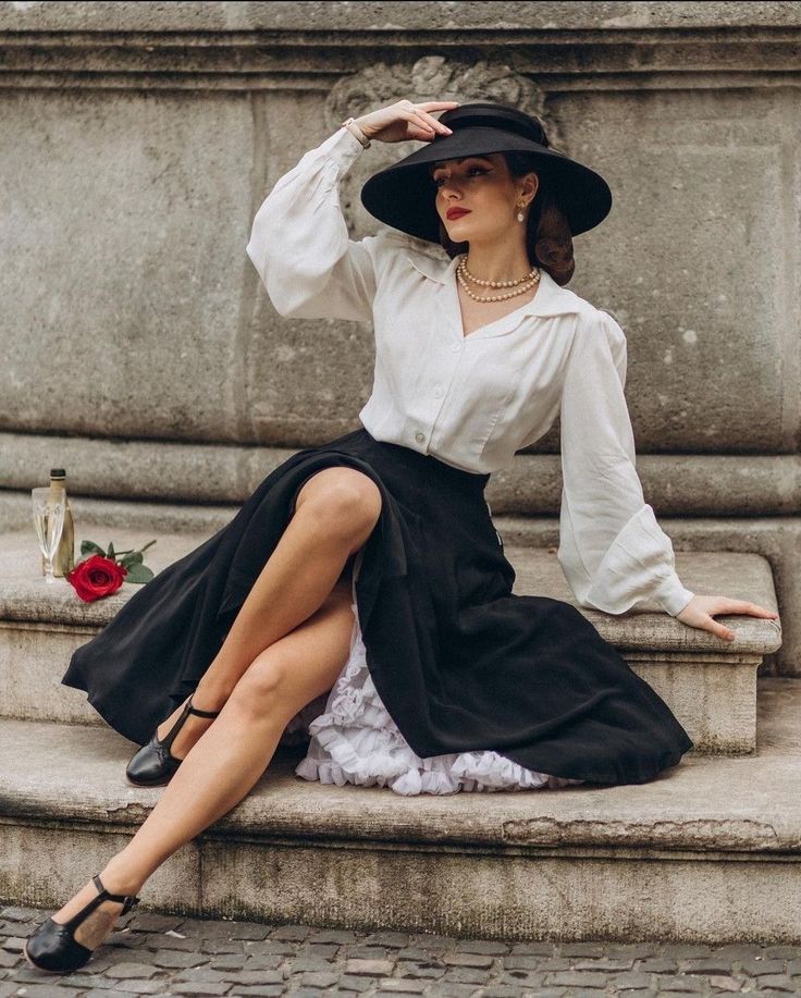 Woman in vintage fashion sitting on stone steps, wearing a black hat and elegant outfit with red rose and champagne nearby. vintage dresses