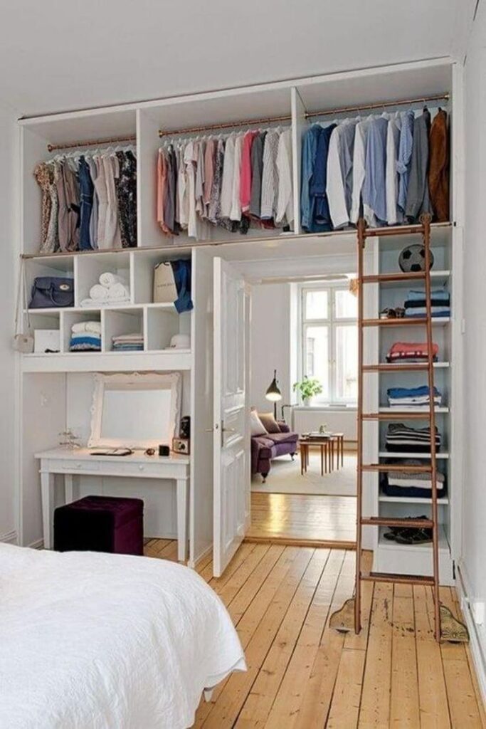Spacious bedroom with open wardrobe, ladder, and neatly organized clothes, leading into a cozy living room with natural light.
