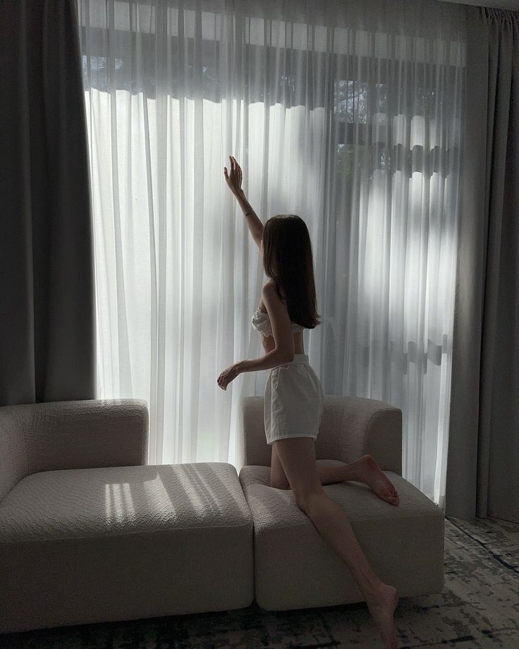 Woman in white outfit reaching towards sheer curtains in softly lit room.