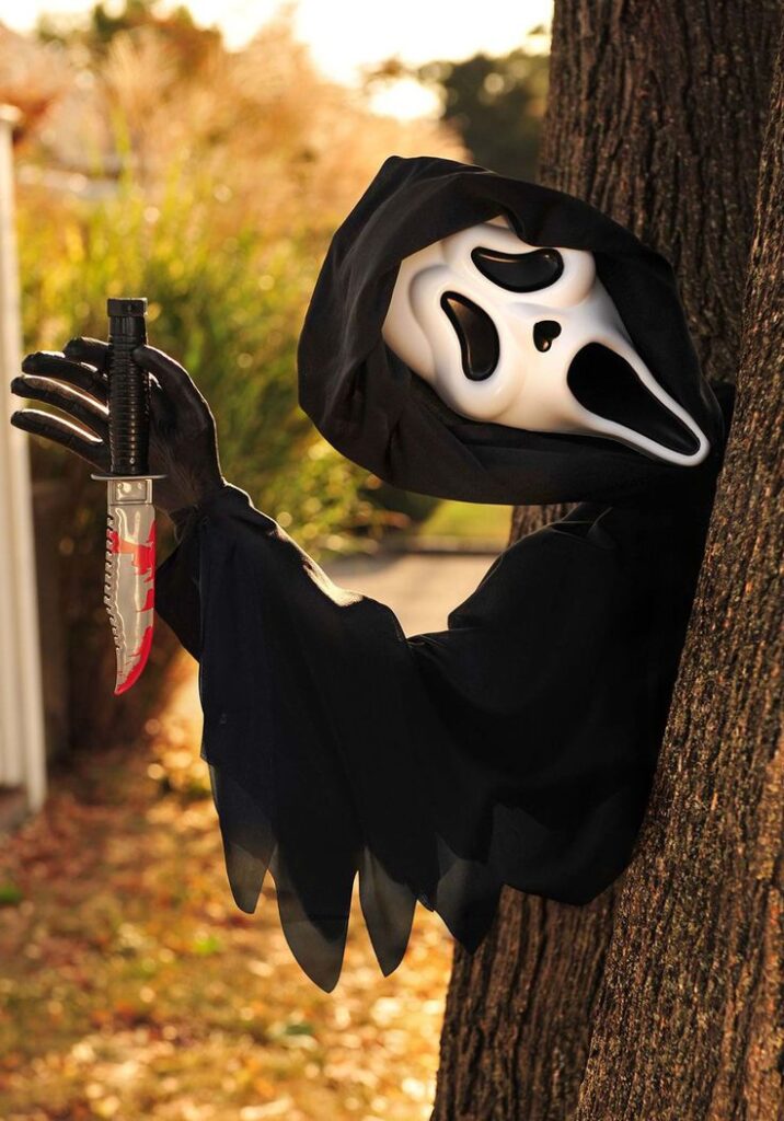 Ghostly figure in black cloak holding bloodstained knife by a tree, wearing a white mask. Spooky Halloween scene.