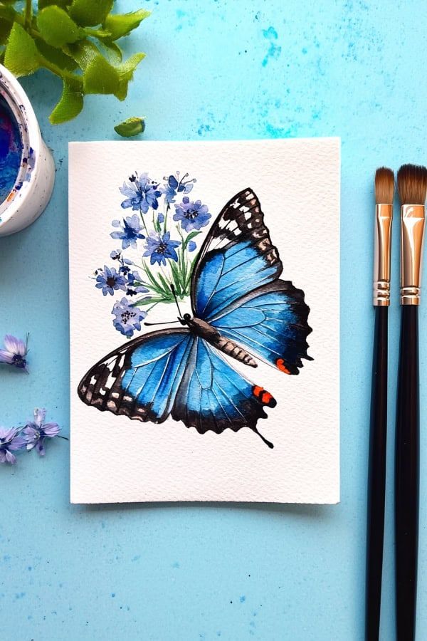 Blue butterfly watercolor painting with flowers on textured paper, set beside paintbrushes on a light blue background.