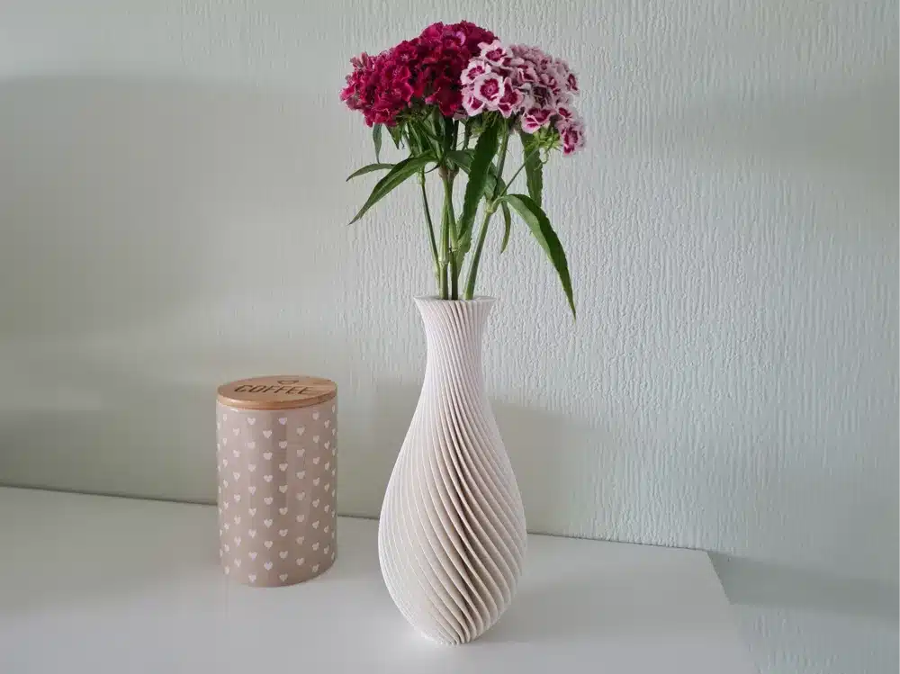 White vase with pink flowers beside a heart-patterned coffee container on a white table against a textured wall.