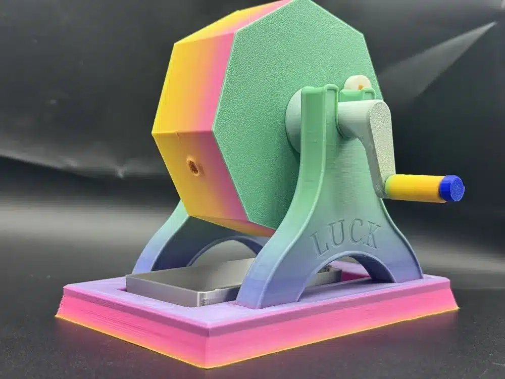 Colorful 3D-printed raffle drum with crank handle on black background, featuring the word LUCK.