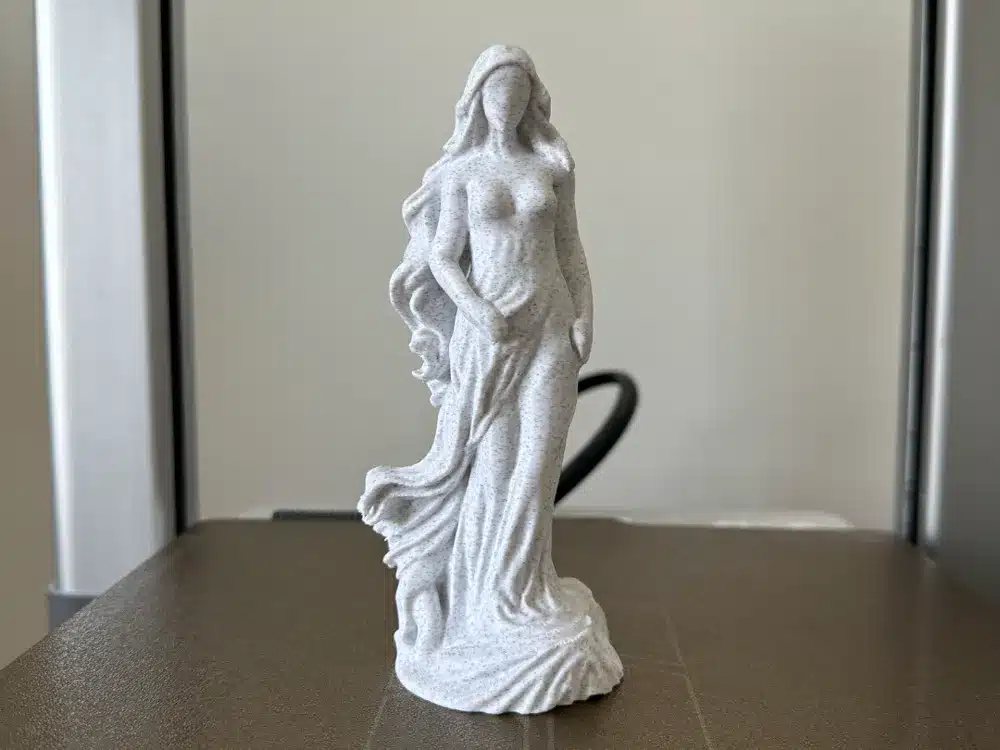 3D printed white statue of a woman with long flowing hair and a draped dress, placed on a table in natural light.