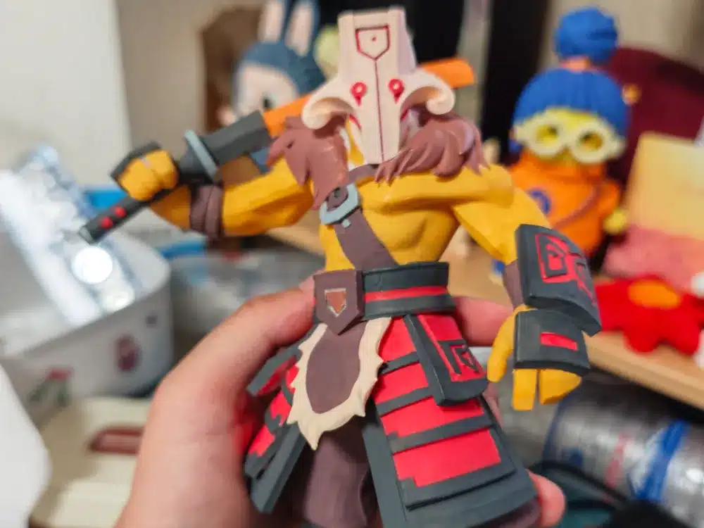 Hand holds action figure in detailed armor, striking a dynamic pose with a sword, colorful shelf background.