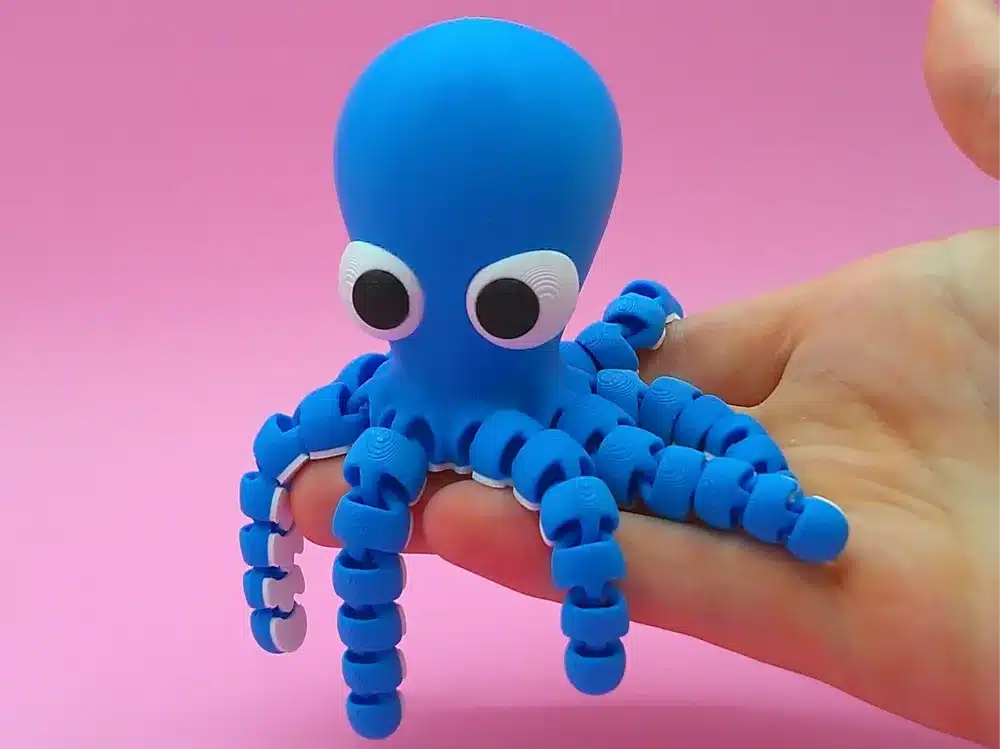 Blue 3D-printed octopus toy with big eyes, held in hand against a pink background.