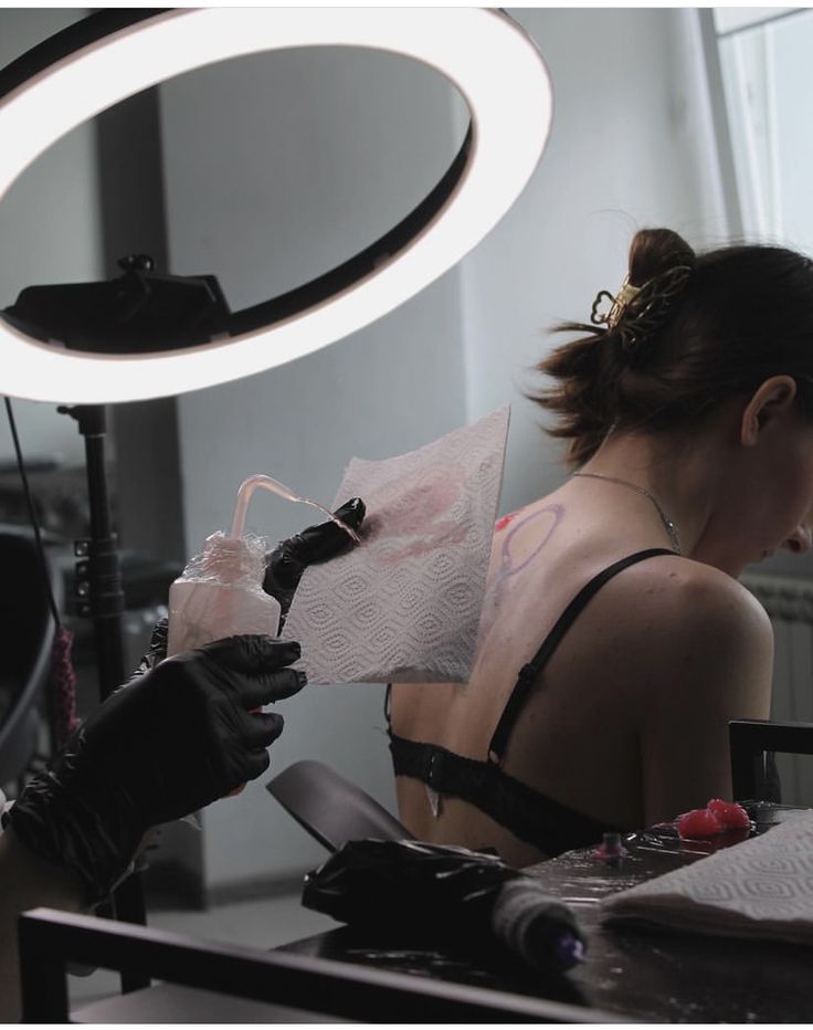 Tattoo artist working on a woman's back under a ring light, using ink and paper towels.