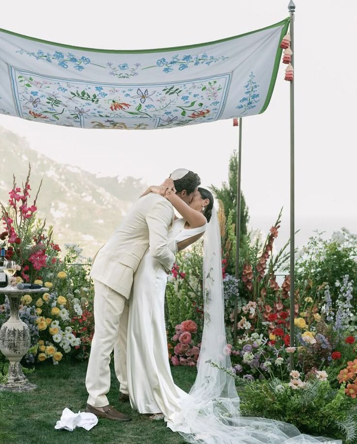 Bride and groom kiss under floral wedding canopy, surrounded by vibrant flowers and scenic landscape.