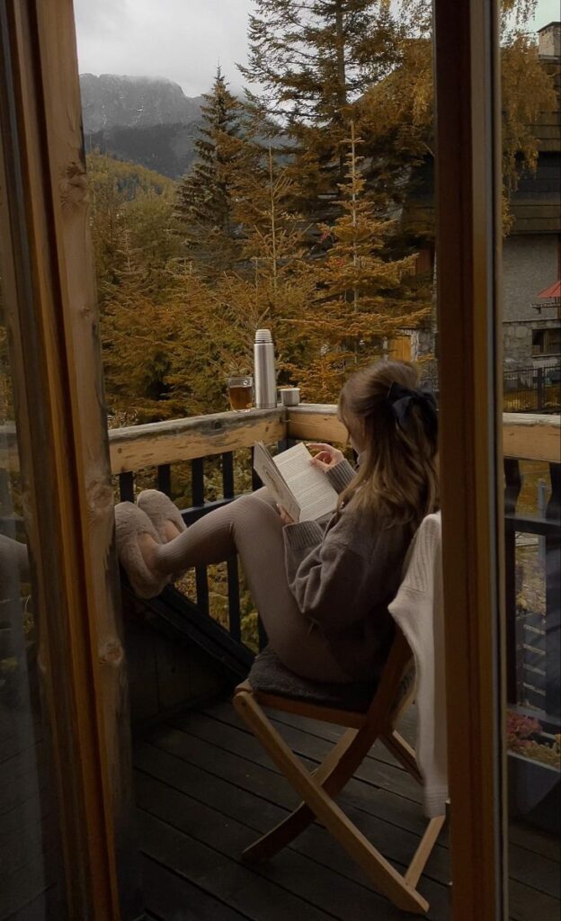 Cozy autumn balcony reading: woman relaxes with book, tea, and mountain view.