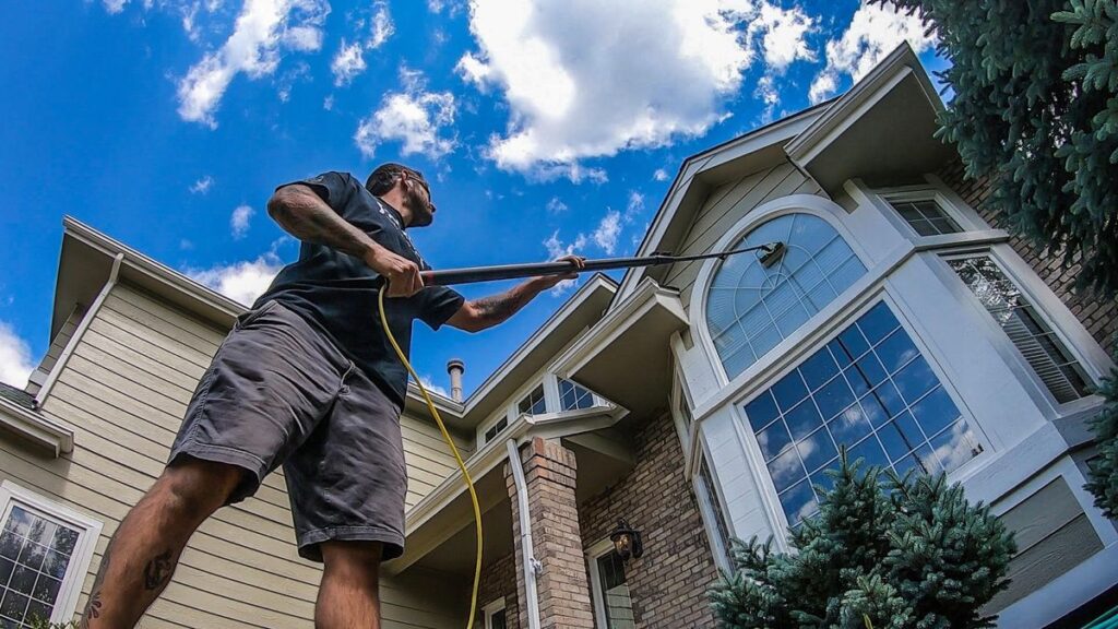 Man cleaning house windows with a pressure washer under a blue sky, emphasizing home maintenance and care.