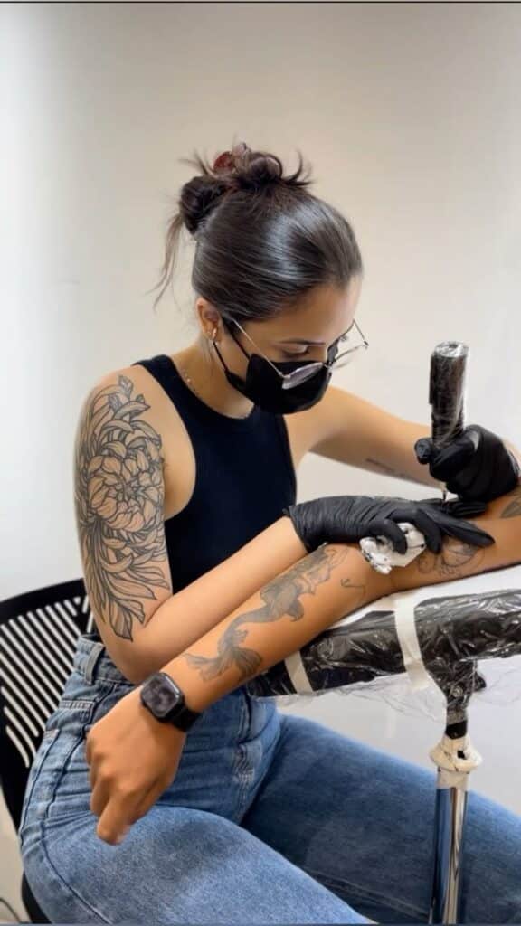 Tattoo artist in black top creating intricate arm design while wearing a mask and gloves.