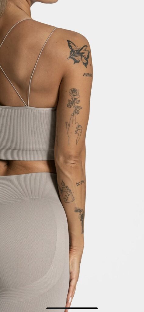 Woman in beige top and leggings, showcasing tattoos on arm and back, including a butterfly and rose design. Arm tattoos