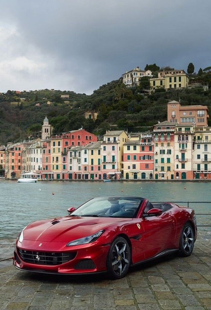 Red convertible parked by waterfront with colorful hillside buildings in the background.