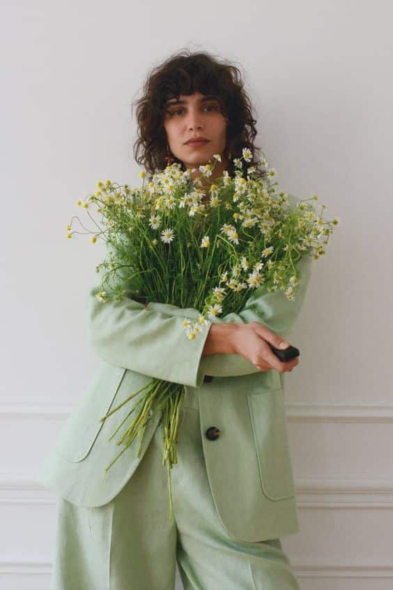 Person in a pastel green suit holding a bouquet of daisies against a white background.