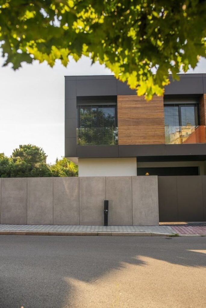 Modern house exterior with wooden accents and concrete fence, shaded by lush green trees.
