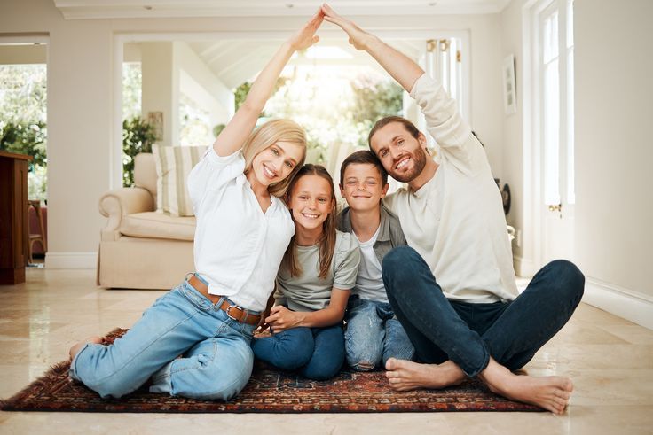 Happy family sitting on the floor forming a house shape with their arms, smiling in a bright, cozy living room.