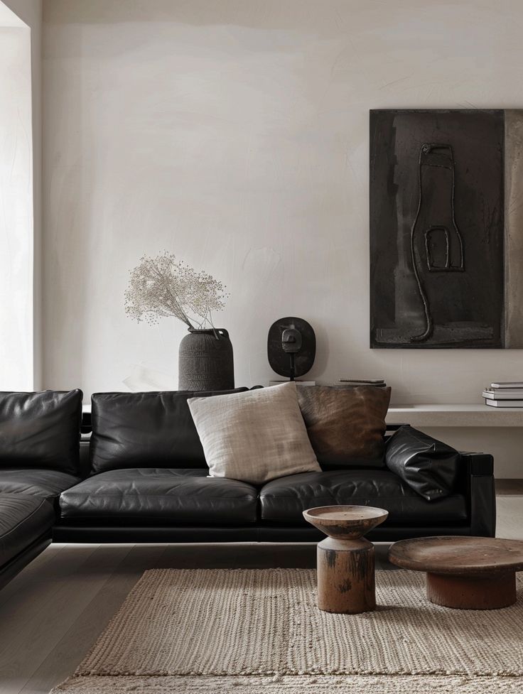 Modern living room with black leather sofa, textured pillows, rustic wooden tables, and minimal art on a neutral wall. Industrial minimalism