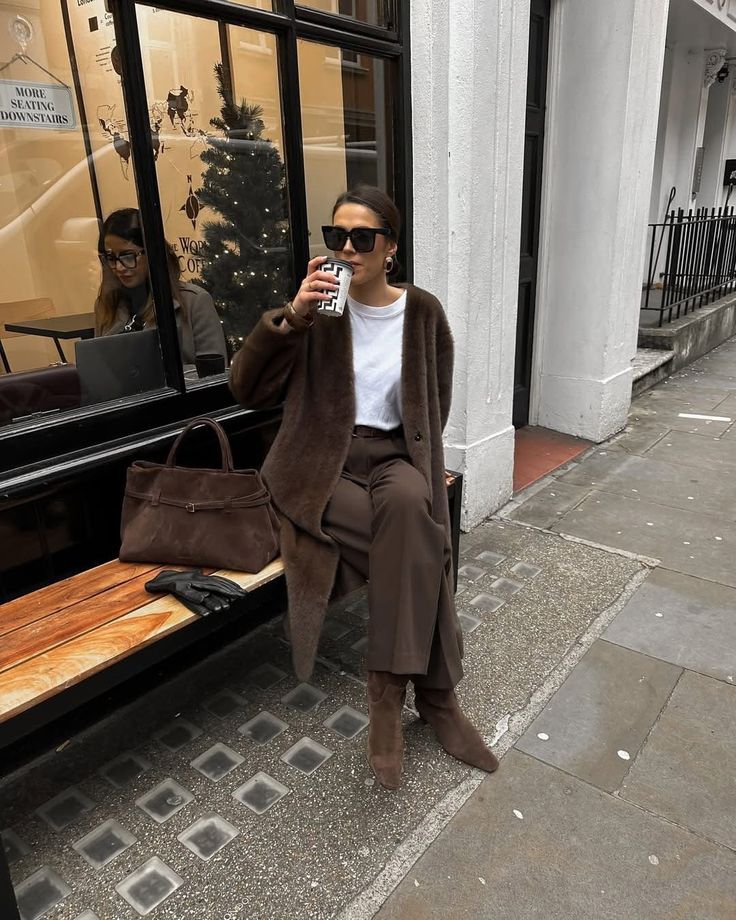Woman in a brown coat sitting on a bench, drinking coffee outside a cafe, with a holiday window display in the background.
