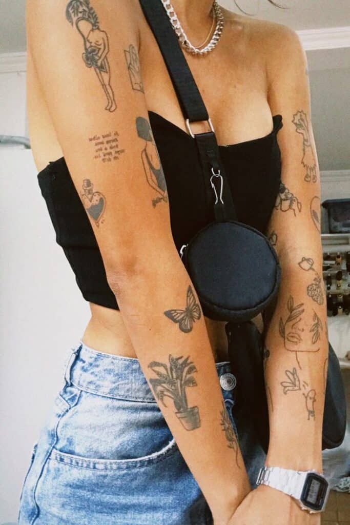 Person in black top and denim shorts showcasing various arm tattoos, wearing a small black crossbody bag.