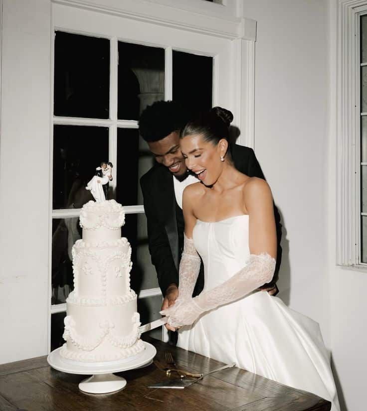 Bride and groom joyfully cutting elegant white wedding cake with topper in bright room.