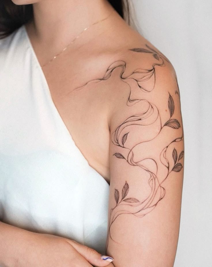 Elegant floral tattoo design on woman's shoulder in a white outfit, showcasing intricate leaf patterns and swirls.