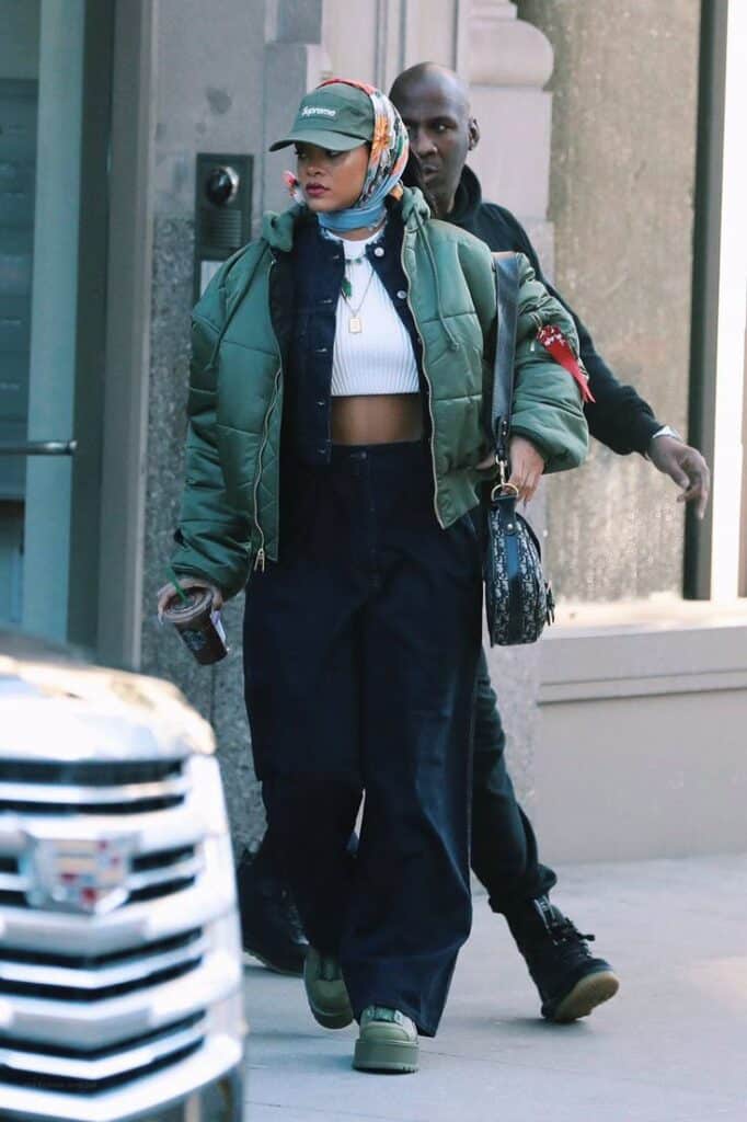 Fashionable woman in green jacket, wide pants, and cap, walking in urban setting with shopping bag and phone.